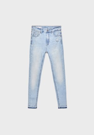 Femme portant un haut ajusté boutonné marron foncé, un jean bleu clair, des bottes marron foncé montant jusqu'aux genoux, avec un sac à épaule marron foncé.