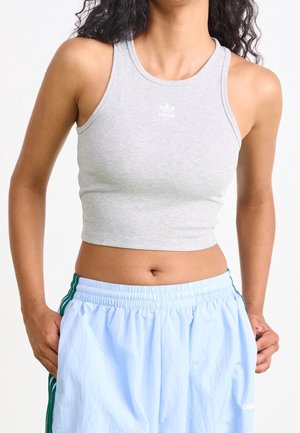 Mujer con un top corto de canalé gris de Adidas y pantalones deportivos azul claro de Adidas con rayas verdes, con las manos en los bolsillos.