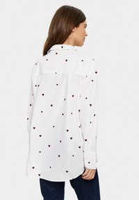 Camicia bianca a maniche lunghe con piccoli cuori ricamati in bordeaux, colletto con bottoni e orlo arrotondato. Vista posteriore mostrata.