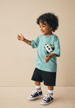 Jeune enfant aux cheveux bouclés jouant avec des bulles, portant un t-shirt bleu à motif de fusée, un short noir, des chaussettes blanches et des baskets noires.