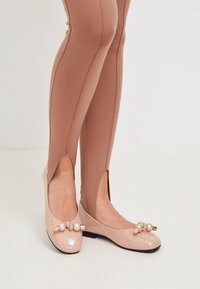 Ballerines nudes en vernis avec un bout arrondi, ornées de perles décoratives et d'un détail en nœud. Portées avec des leggings ajustés en côtes de couleur marron clair.