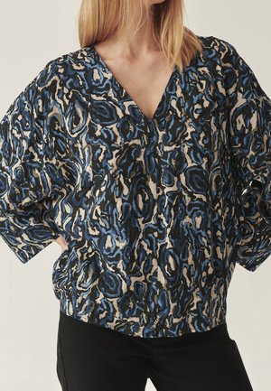 Frau trägt eine locker sitzende V-Ausschnitt-Bluse mit einem abstrakten Muster in Blau, Schwarz und Beige sowie schwarze Hosen.