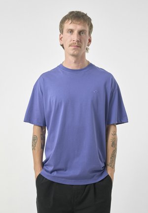 Purple crew neck t-shirt made of cotton, featuring short sleeves, a loose fit, and a small logo on the chest, paired with black pants.
