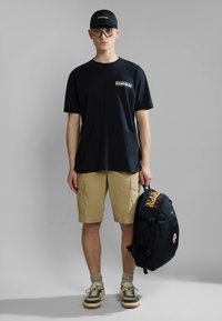 Joven con gorra negra, camiseta negra de gran tamaño, pantalones cortos caqui, zapatillas y gafas, sosteniendo una mochila negra con parches de logo.
