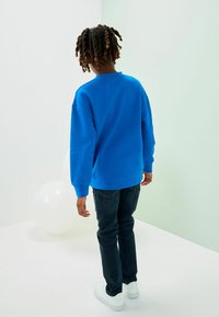 Felpa blu con spalle abbassate, polsini e orlo a coste; abbinata a jeans scuri e sneakers bianche, vista da dietro.