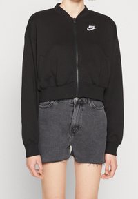 Černá zkrácená mikina se zipem v přední části a žebrovanými manžetami, na levé horní straně je bílá logo Nike. V kombinaci se šedými džíny šortkami.
