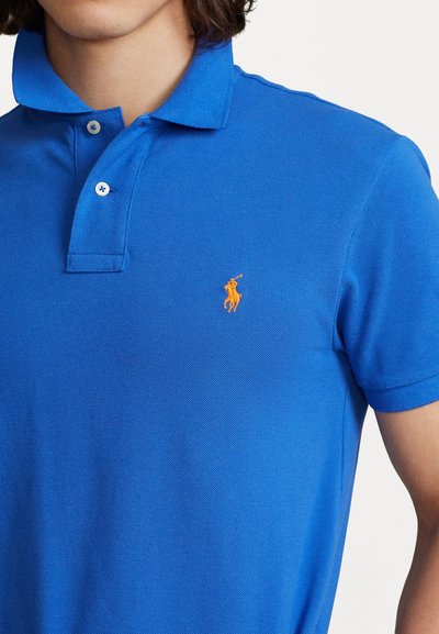 Sinine polo särk tekstureeritud kangast, millel on kolm nuppu ja rinnas väike oranž brodeeritud logo.