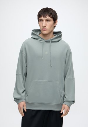 Ung mand med kort mørkt hår iført en lysegrå oversized hættetrøje og sorte bukser, stående mod en ensfarvet hvid baggrund.
