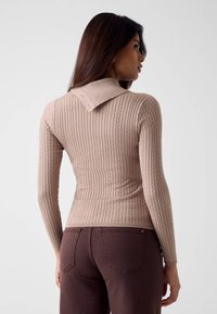 Maglione beige a coste con collo ripiegato e maniche lunghe. Il tessuto sembra strutturato. Abbinato a pantaloni marrone scuro.