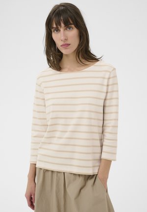 Žena stojaca, oblečená v béžovo-bielom tričku s vodorovnými pruhmi na dlhý rukáv a vo voľných béžových nohaviciach, pravou rukou vrecko.