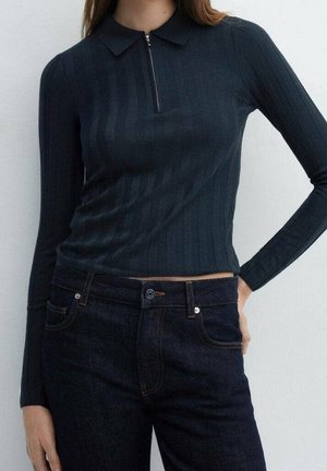 Kvinde iført en tætsiddende, langærmet sort ribbet top med en kort lynlås foran og mørke højttaljede jeans, hånd hvilende på hoften.