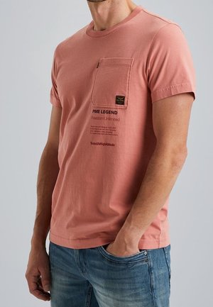 Mężczyzna w koralowo-różowej koszulce z krótkim rękawem i kieszonką na piersi z czarnym napisem, ubrany w niebieskie dżinsy, z ręką w kieszeni.