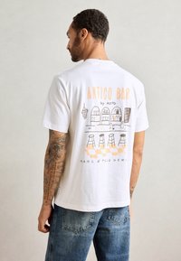 Witte katoenen t-shirt met een grafische print op de achterkant met de tekst "ANTICO BAR" in oranje, met een afbeelding van een barscène met krukken.