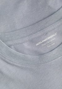 Camiseta de algodón gris con cuello redondo, con una textura suave y costuras visibles, etiquetada "JACK & JONES ORIGINALS", talla grande.