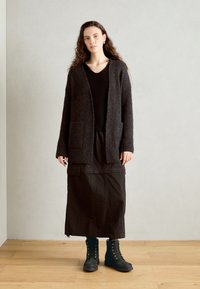 Svart, räfflad kofta med öppen front och två fickor, bärs över en strukturerad svart topp och en lång, mörk kjol. Matchas med svarta snörstövlar.