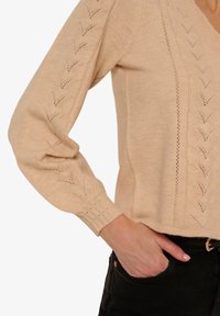 Beżowy sweter z dzianiny z dekoltem w kształcie V, z wzorami liści na rękawach i bokach, teksturowanymi ściągaczami oraz luźnym krojem.