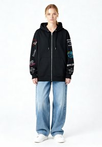 Hanorac negru cu fermoar și glugă, având broderii colorate și text pe mâneci, asortat cu blugi largi, albastru deschis și pantofi albi.