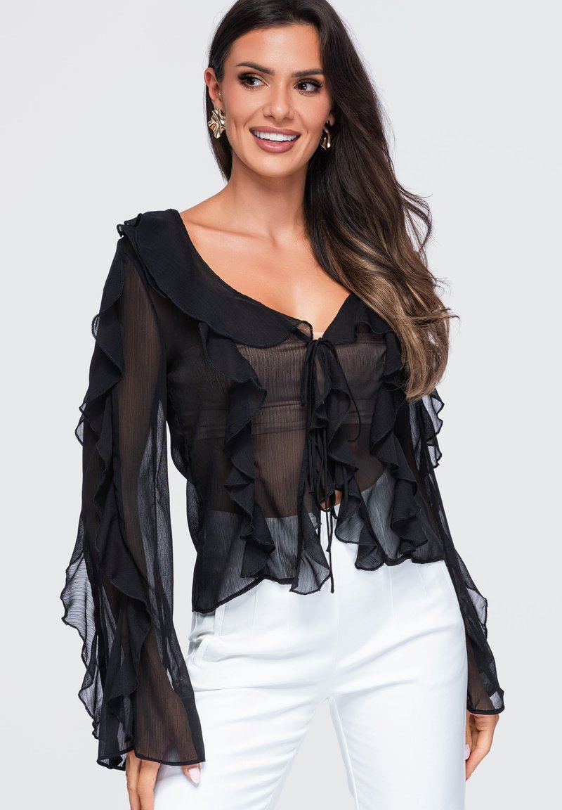 Femme portant un chemisier noir transparent à volants avec un nœud à l'avant et un pantalon blanc, souriante et regardant sur le côté avec de longs cheveux foncés.