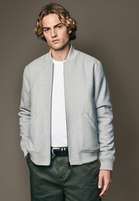 Light gray bomber jacket with ribbed cuffs and waistband, zip front, side pockets, worn over a white t-shirt and olive green pants.