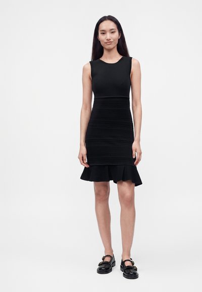 Femme debout portant une robe noire sans manches avec une partie centrale texturée et un ourlet volanté, associée à des chaussures Mary Jane noires.