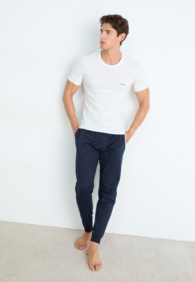 T-shirt blanche à manches courtes avec un petit logo "BOSS" noir sur la poitrine, associé à un pantalon tapered bleu marine. Pieds nus sur un tapis clair.