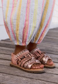 Sandali in camoscio rosa con dettagli metallici, caratterizzati da cinturini intrecciati, design traforati e suola piatta, abbinati a pantaloni leggeri a righe.