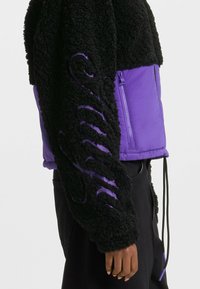 Giacca nera e viola con parte superiore in pile soffice e parte inferiore in tessuto liscio. Presenta una tasca con zip e un cordino all'orlo.