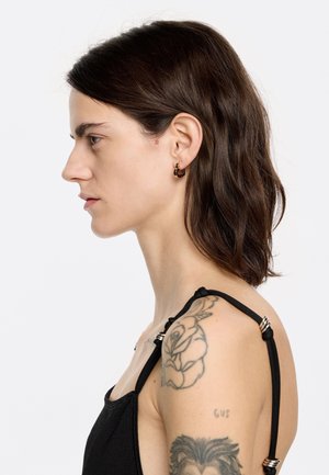 Zelta apli auskari ar savijumiem melniem akcentiem, demonstrējot glītu dizainu. Modelis valkā melnu apģērbu, un uz pleca ir redzams tetovējums.