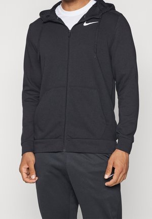 Mężczyzna ubrany w czarną bluzę Nike z kapturem na zamek i pasujące czarne spodnie dresowe Nike, ręce swobodnie opuszczone wzdłuż ciała.