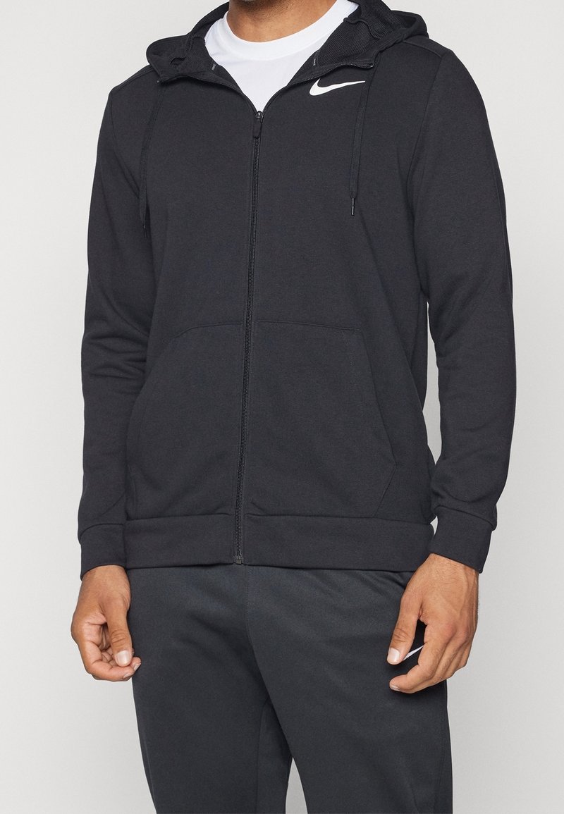 Homme portant un sweat à capuche zippé noir Nike avec capuche et un pantalon de jogging noir assorti Nike, mains détendues le long du corps.