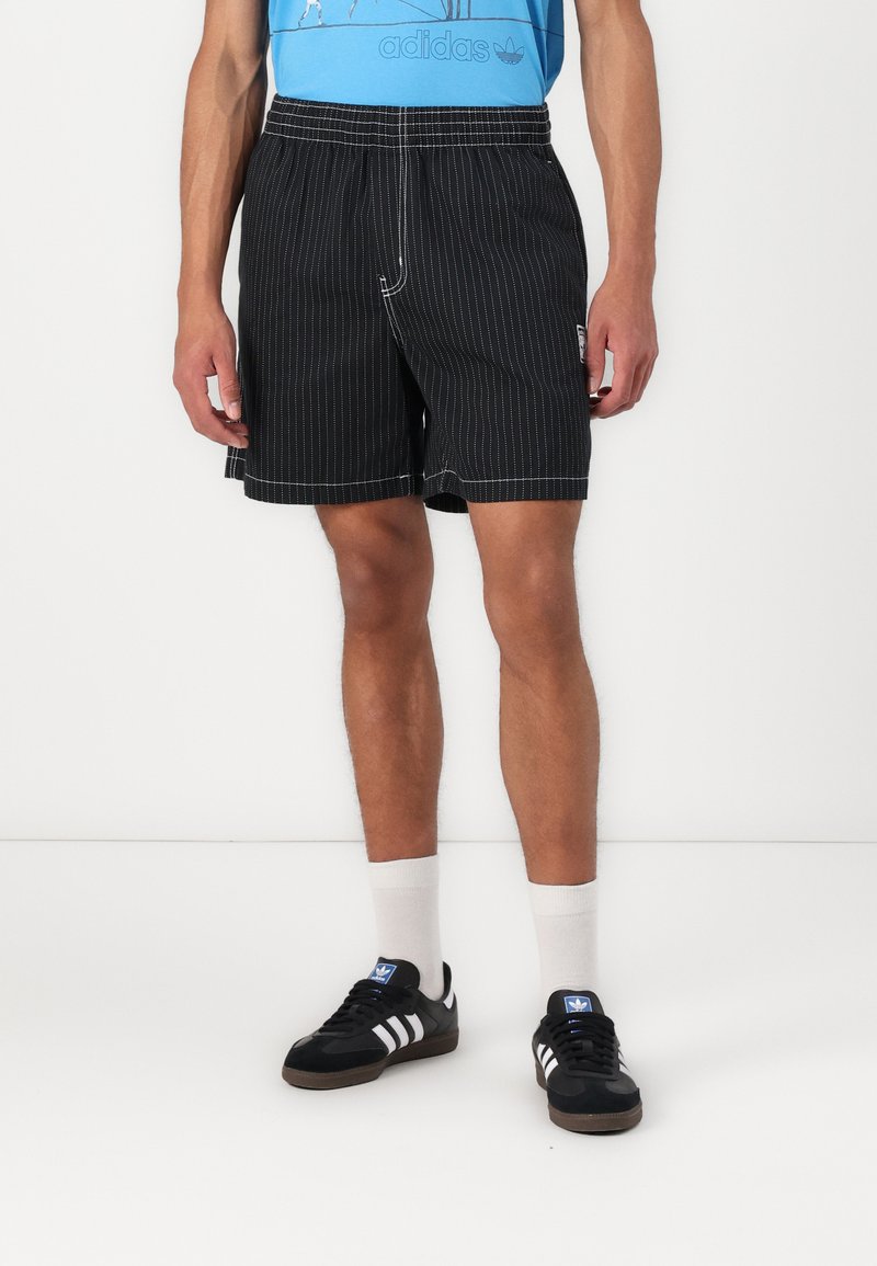 Fekete csíkos rövidnadrág, elasztikus derékkal és elülső zsebekkel, fekete Adidas tornacipővel és fehér, térdhosszú zoknival párosítva.