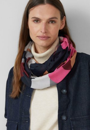Kobieta o długich brązowych włosach nosi wielokolorowy szalik na kremowym golfie i ciemnoniebieskiej, zapinanej na guziki kurtce na jednolitym szarym tle.