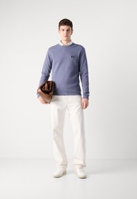 Lyseblå bomullsgensar med rund hals, iført over en hvit skjorte med krage, kombinert med hvite denimbukser og beige sneakers. Holder en brun skinnveske.