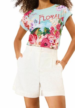 Femme portant un t-shirt bleu clair à imprimé floral avec le mot "Floral" et un short blanc taille haute avec poches, les mains dans les poches.