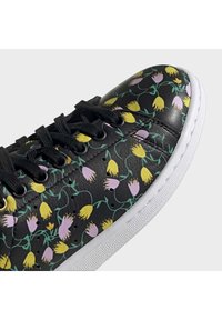 Černá teniska s černými tkaničkami, zdobená žlutými a světle fialovými květinovými vzory a zelenými stonky, se světlou texturovanou podrážkou.