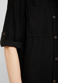 Schwarzes kurzärmliges Hemd mit einer einzelnen Brusttasche, aufgeschlagenen Ärmeln und Knopfdetails. Glattes Gewebe mit subtiler Textur und Nahtdetails.