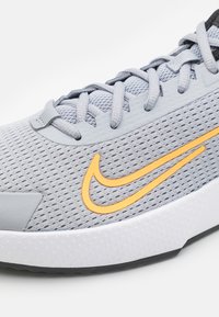 Scarpa sportiva grigia con tomaia in rete, logo Nike arancione, dettagli testurizzati e una suola bianca resistente. I lacci sono grigi e funzionali.