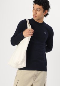 Ναυτικό μπλε πλεκτή ζακέτα με μακριά μανίκια και στρογγυλή λαιμόκοψη, σε συνδυασμό με μια κρεμ καμβά τσάντα. Διακριτική λεπτομέρεια λογότυπου στη ζακέτα.