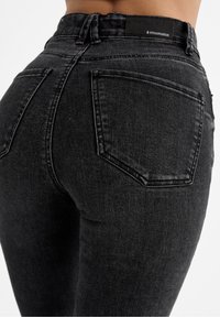 Jeans de mezclilla negra con una cintura alta, ajuste slim y diseño de cinco bolsillos. La tela tiene un acabado ligeramente texturizado y detalles de costura sutiles.