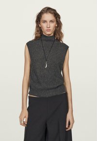 Dunkelgrauer ärmelloser Rollkragenpullover aus weichem Strickstoff, mit tailliertem Schnitt und strukturierter Oberfläche. Kombiniert mit schmal geschnittenen schwarzen Hosen.