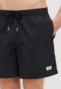 Czarne kąpielówki wykonane z lekkiego materiału, z elastycznym paskiem w talii i sznurkiem, kieszeniami po bokach oraz małą metką z materiału.