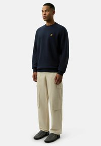 Granatowy sweatshirt z małym złotym logo, beżowe spodnie cargo z bocznymi kieszeniami i szare sneakersy w minimalistycznym otoczeniu.