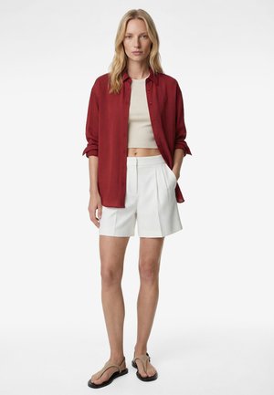 Kvinde iført en rød skjorte med knapper over en beige crop top, hvide plisserede shorts og beige sandaler, stående med den ene hånd i lommen.