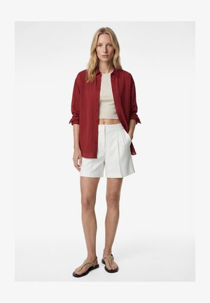 Mujer con camisa roja de botones sobre un crop top beige, pantalones cortos blancos plisados y sandalias beige, de pie con una mano en el bolsillo.