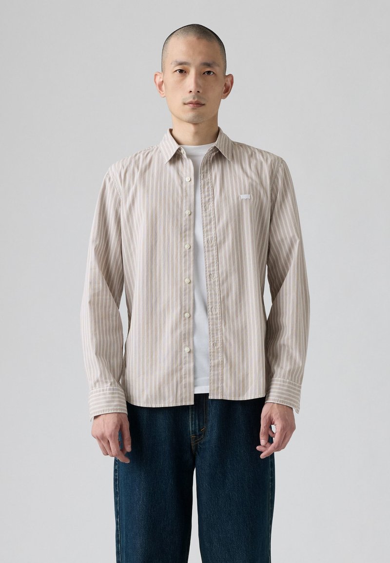 Camicia button-up a righe in beige chiaro con sottili strisce marroni. Presenta un colletto classico, maniche lunghe e un logo sul petto. Indossata sopra una maglietta bianca.