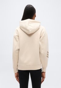 Μπεζ φούτερ με κουκούλα και χαλαρή εφαρμογή, με ριμπ μανσέτες και τελείωμα, και τρεις οριζόντιες ρίγες στο αριστερό μανίκι. Ομαλή υφή.