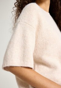 Nærbillede af en person iført en blød, lysebeige kortærmet strikket sweater, der viser skulder, arm og delvist nakke med krøllet hår.