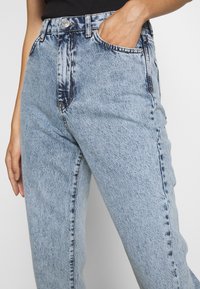 Ljusblå denimjeans med hög midja, rak ben, synlig sömnad och klassisk femficksdesign. En hand är placerad i fickan.