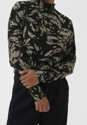 Personne portant un chemisier noir à col montant avec des motifs abstraits gris en forme de plumes, associé à un pantalon noir taille haute, bras croisés.