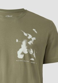 T-shirt vert olive avec imprimé floral abstrait blanc et texte en cursive "less is more - less is bore synergize" sur le devant.